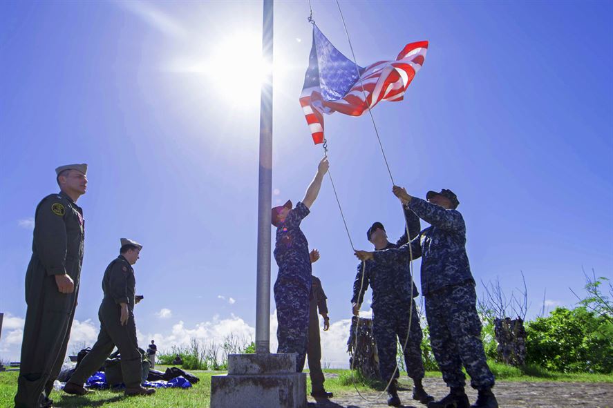 Three U.S. sailors in camouflage raise an American flag on a flagpole with the sun shining behind them. 
