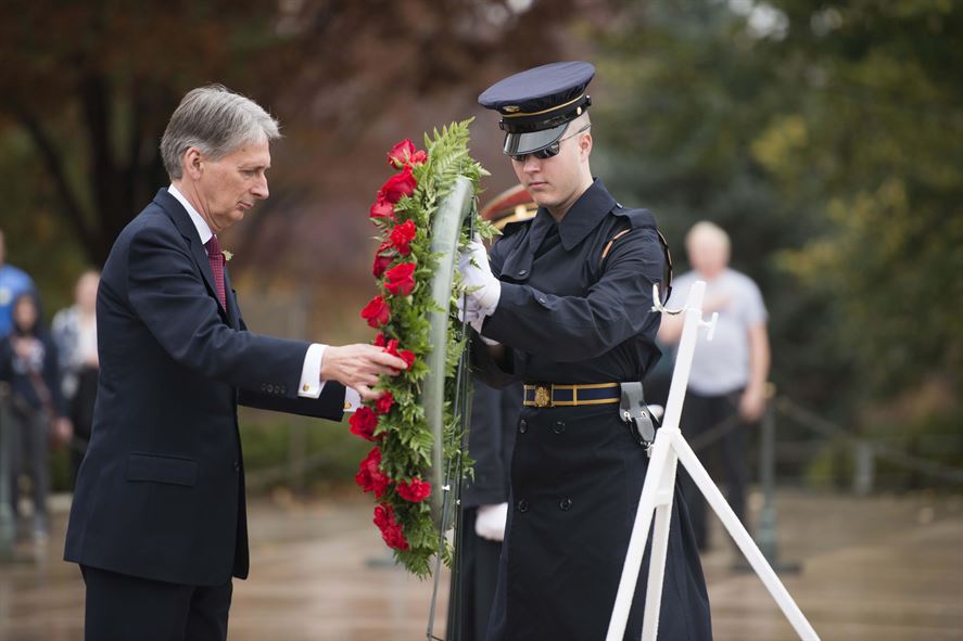 British Foreign Secretary Philip Hammond lays a large wreath with green leaves and red flowers with assistance from a young male marine.