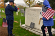 Uniformed military soluting a grave while another person holds an American flag.