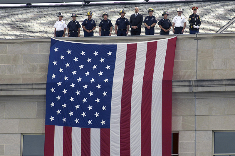 Representatives of police and firefighting units who were the first to respond to the 9/11 terrorist attack on the Pentagon, stand at the top of a large American flag suspended from the roof of the Pentagon.