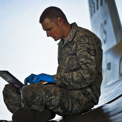 A service member sits cross-legged on the ground using a laptop.