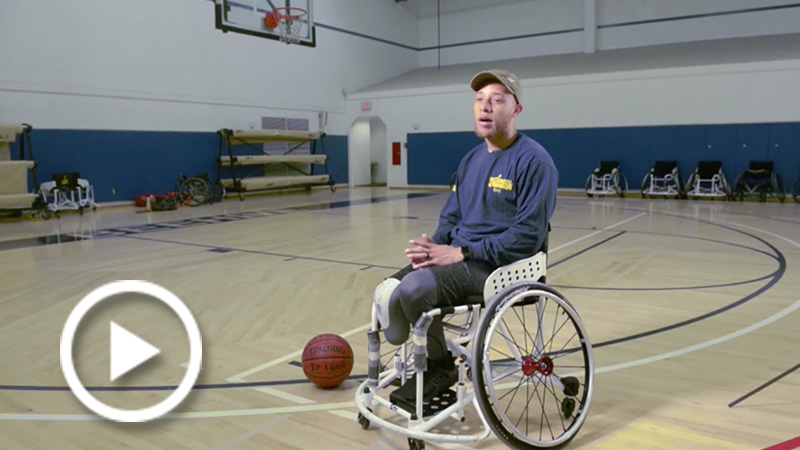 Petty Officer AJ Thomas sits in a wheelchair being interviewed on a basketball court.
